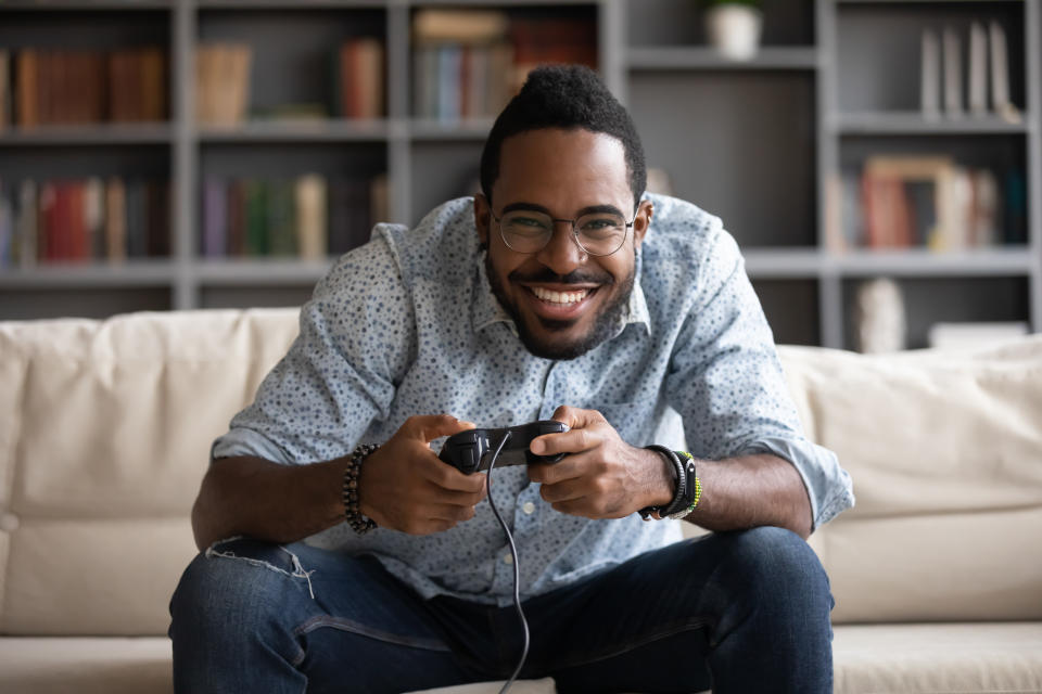 Overjoyed African American millennial man relax on couch in living room play video game using joystick gamepad, happy biracial young male gamer rest at home engaged in digital virtual activity