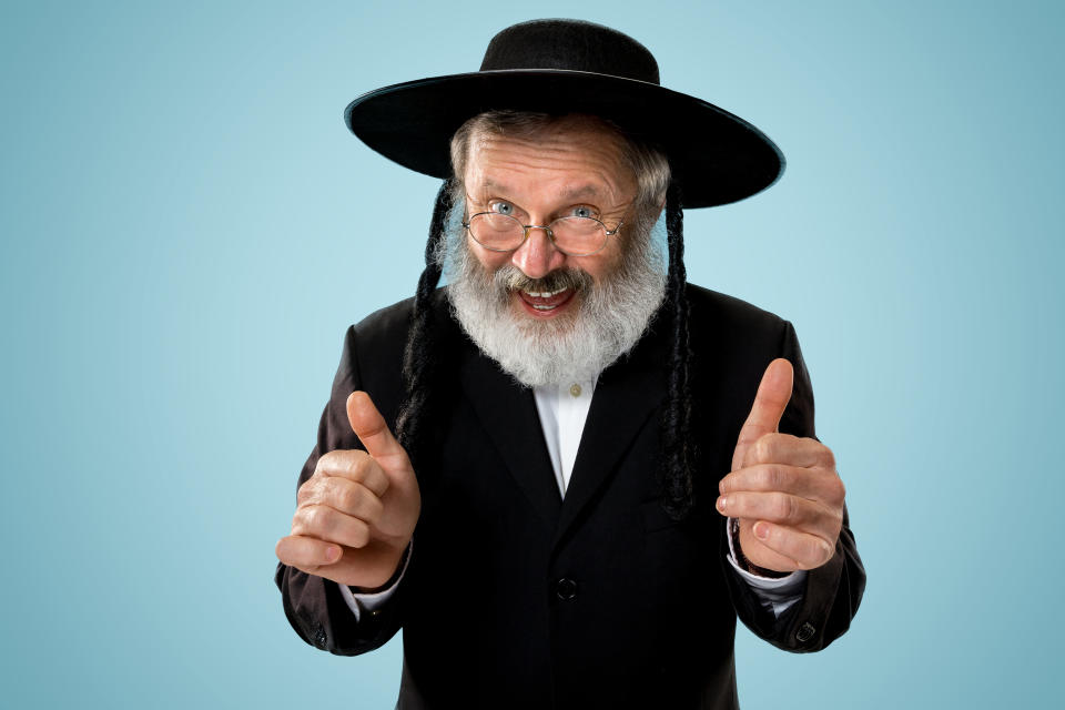 An older man with a beard and sidelocks, wearing a black hat and suit, smiles and gives two thumbs up against a plain background