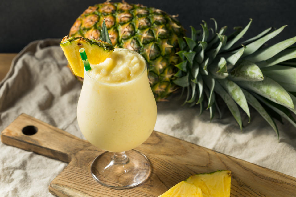 A piña colada in a glass garnished with a pineapple slice and a straw, with a whole pineapple in the background on a wooden board