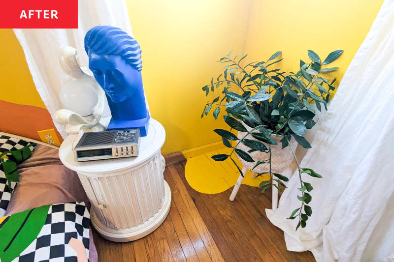 House plant in white wicker plant stand next to yellow painted floor decal in newly painted yellow bedroom. Pillar beside bed holds sculptures and vintage alarm clock.