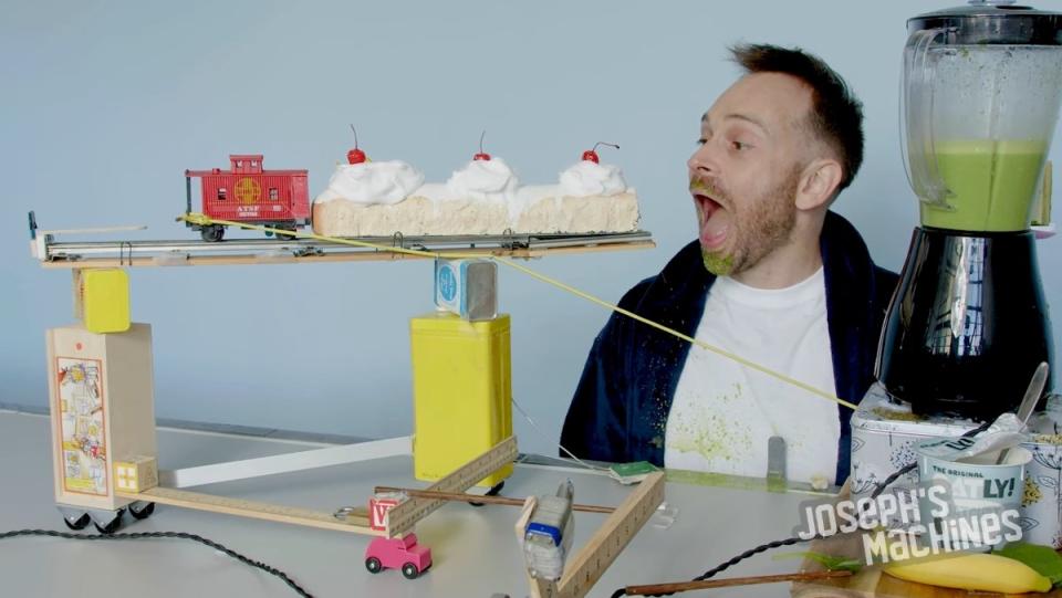 A man sits while a conveyer belt passes with a blender of green smoothie and a large pastry