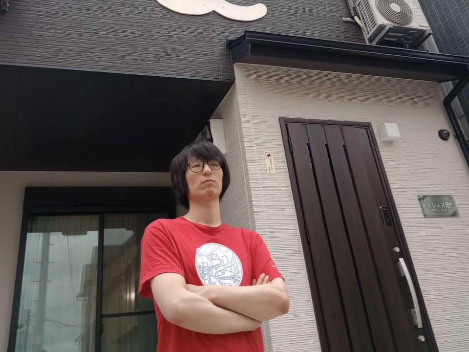 Jin Gujin standing outside a small apartment while folding his arms and wearing a red T-shirt with a white circle on the chest.