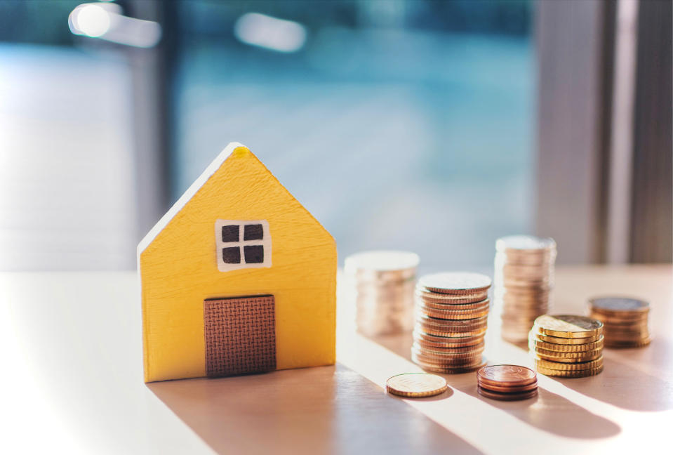 A small yellow wooden house sits on a table next to several stacks of coins of varying heights