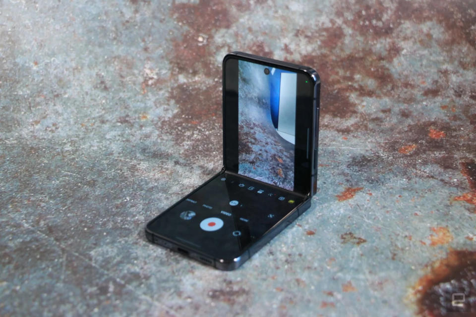 Foto del Samsung Z Flip 5 sentado sobre una superficie que parece metal rugoso con manchas oxidadas.  El teléfono está parcialmente abierto con la vista de la cámara visible en la mitad superior de la pantalla.