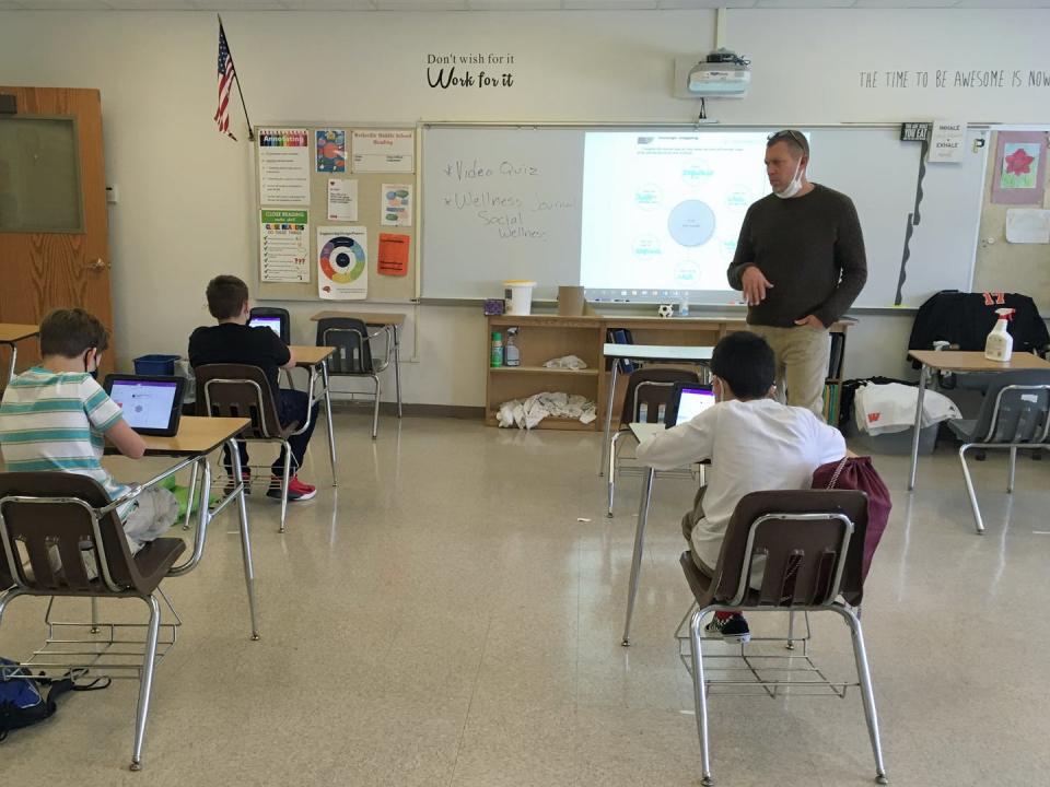 A teacher wearing a mask conducts his class while his students sit using iPads.