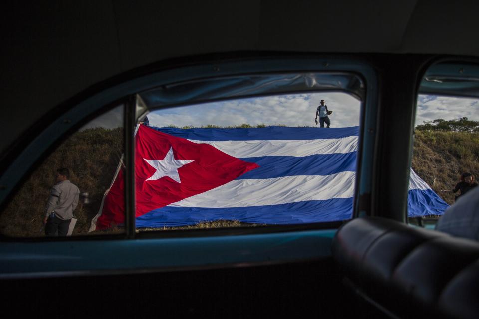 FILE - In this Dec. 2, 2016 file photo, a Cuban flag covers the side of the road where people wait for the funeral procession of Fidel Castro on the outskirts of Las Tunas, Cuba. Castro's ashes crossed Cuba from Havana to their final resting place in the eastern city of Santiago, passing through the small towns and cities where his rebel army fought its way to power nearly 60 years ago.(AP Photo/Desmond Boylan, File)