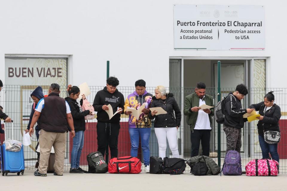 A line of people holding documents, with bags on the ground, outside a white building