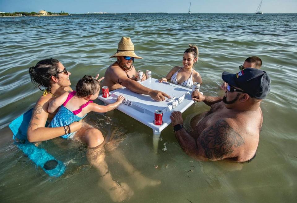 Playing dominoes, Lizzy Reyes holding Sophia Acosta, 2, Ronny Chavez, Dariana Alvares, Magdiel Figueroa and Emilio Reyes, clockwise, cool off in the water along the Rickenbacker Causeway in Miami on Tuesday, June 30, 2020. Miami-Dade County recorded a record-breaking temperature of 98 degrees on Tuesday, according to the National Weather Service.