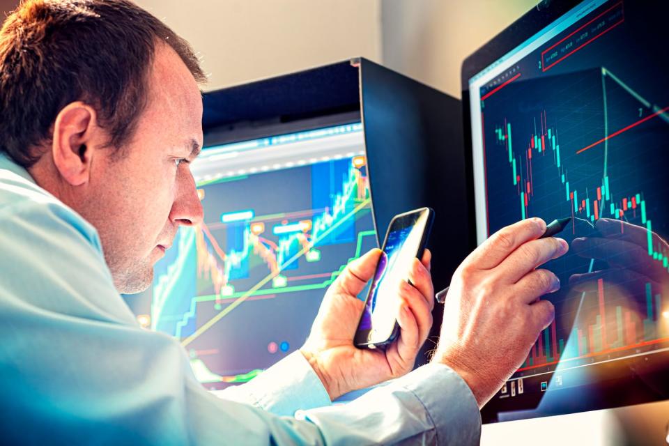 An asset manager uses a pen and a smartphone to analyze a stock chart displayed on a computer monitor.