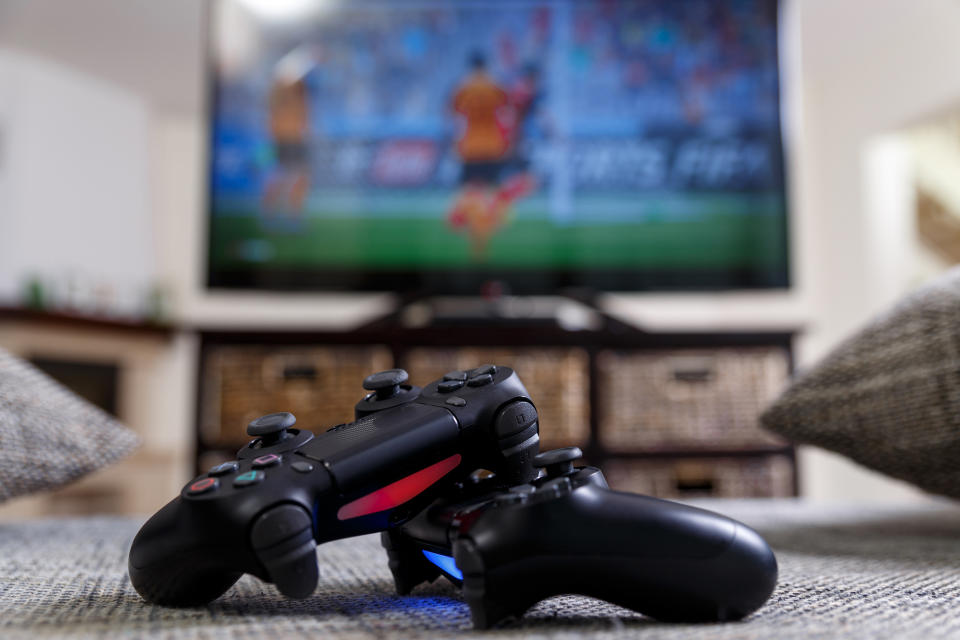 Game console controllers lying on the floor in a living room with a TV turned on in the background.