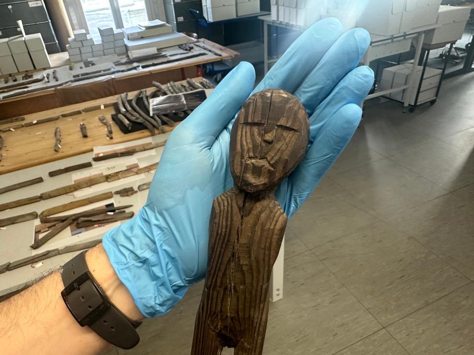 A gloved hand holding a wooden human figure with eyes, a nose, and a mouth carved into its face and no arms.