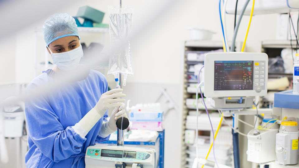 Picture of a nurse, with a face mask on administering medicine through a drip in a hospital, surrounded by medical equipment.