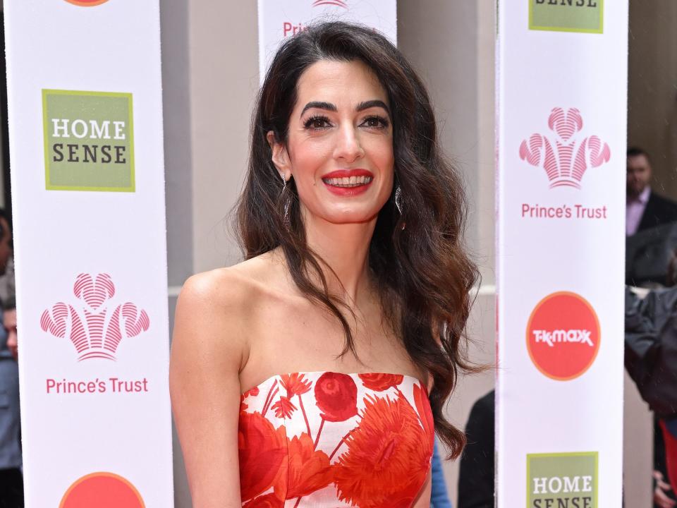 Amal Clooney wears a white dress decorated in a red floral pattern in front of white signs with logos and text saying, "Home Sense," "TJ Maxx," and "Prince's Trust"