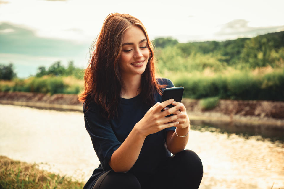 Happy smiling young woman sitting outdoors close to a river looking towards her mobile phone, writing messages with a bright happy smile. Real People Outdoor Youth Social Media Lifestyle.