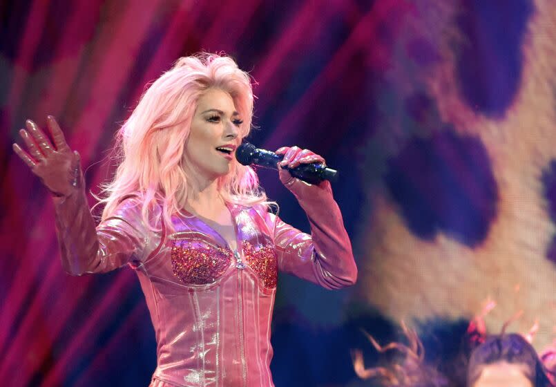 Shania Twain is performing on stage with a microphone to her mouth, dressed in a pink leather cowboy jumpsuit
