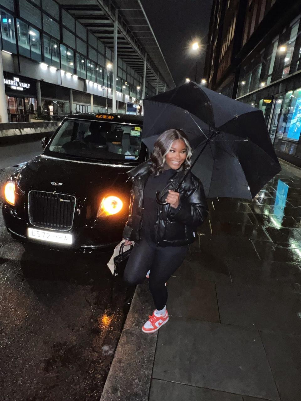 Gladys Nkengasong wears all black and holds an umbrella in front of a black British taxicab.