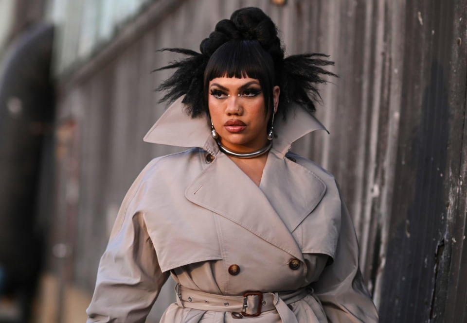 Person with dramatic eye makeup and a high-collared trench coat styled with unique hairdo