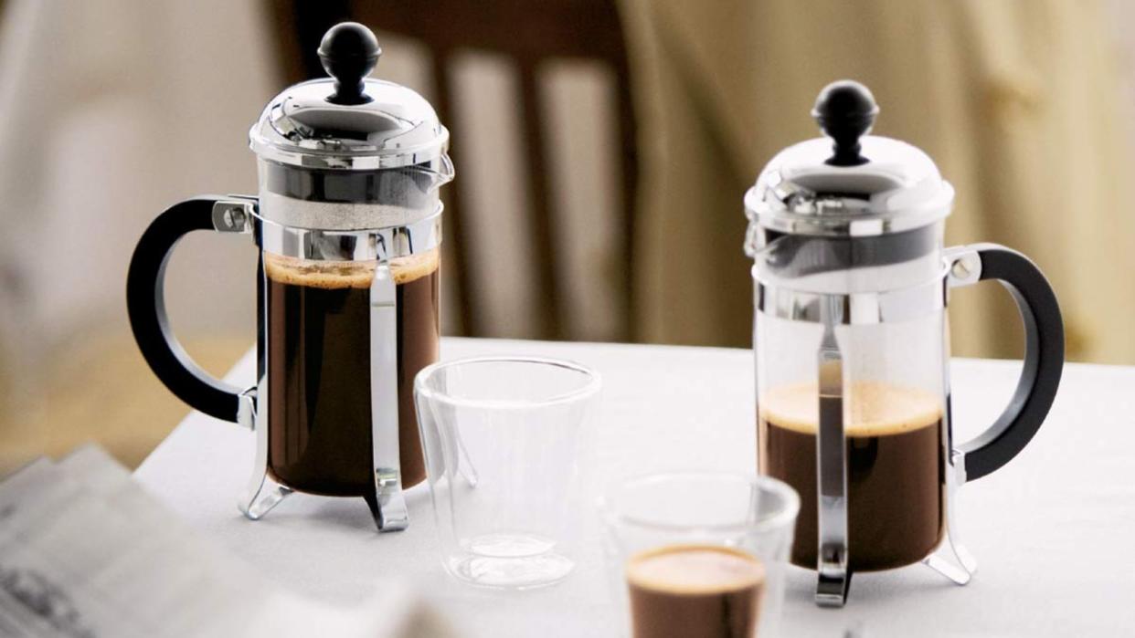  Two Bodum Chambord French presses on a table with a cup of coffee in front of them. 