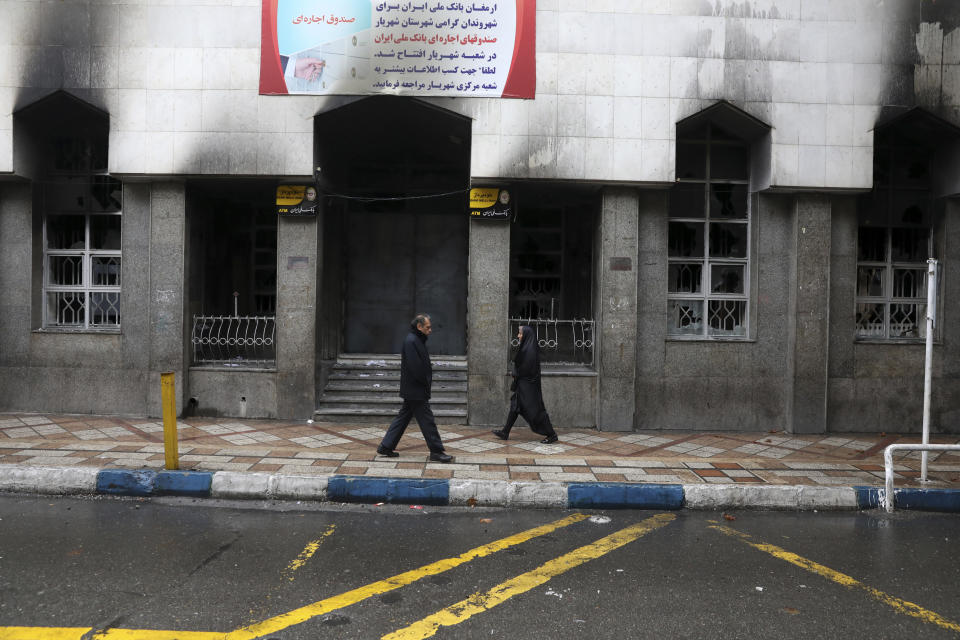 People walk past a bank that was burned during recent protests, in Shahriar, Iran, some 40 kilometers (25 miles) southwest of the capital, Tehran, Wednesday, Nov. 20, 2019. Protests over government-set gasoline prices rising struck at least 100 cities and towns, spiraling into violence that saw banks, stores and police stations attacked and burned. (AP Photo/Vahid Salemi)