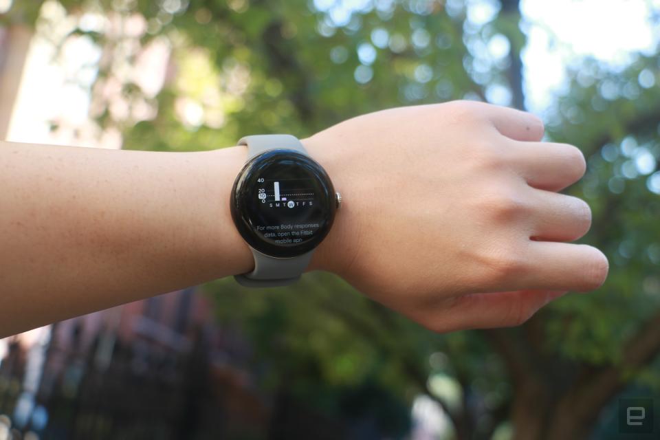 The Pixel Watch 2 worn on a wrist held in mid-air, with sunlight streaming through leaves in the background.
