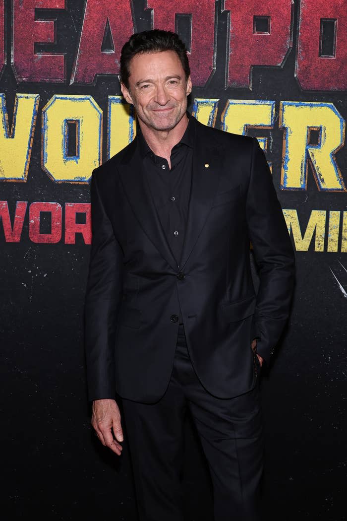 Hugh Jackman at a film event, wearing a black suit and smiling while standing in front of a "Deadpool 3" promotional background