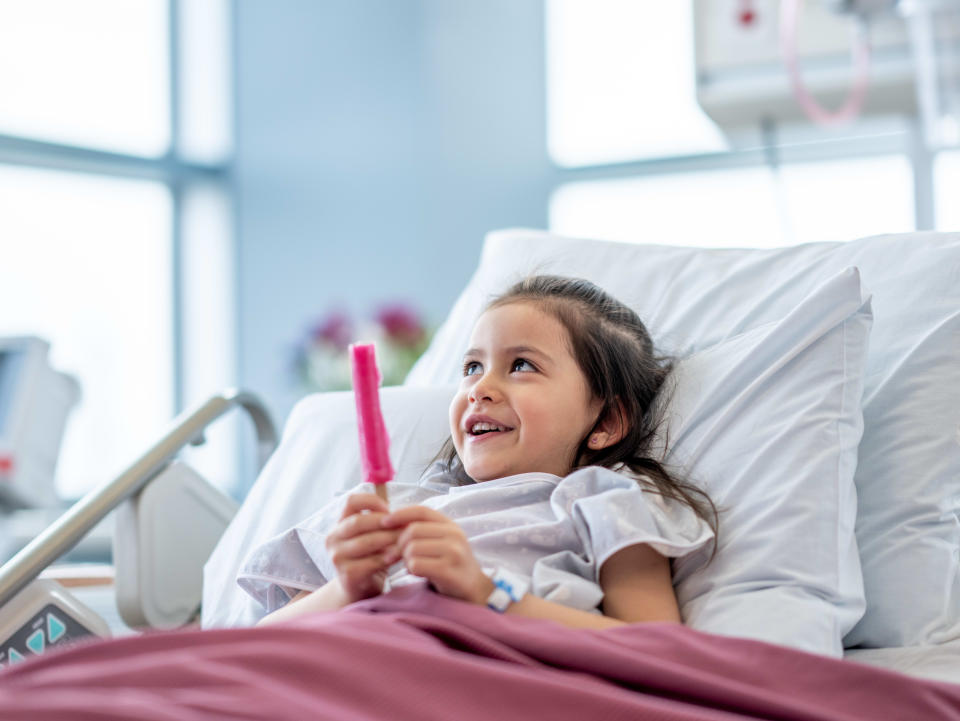 A sweet little girl sits up in her hospital bed after surgery as she eats a popsicle.  She is wearing a hospital gown and tucked warmly under the blankets.