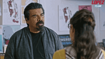 George Lopez talking to a woman in glasses at an indoor location, with posters on the wall and "Lopez vs. Tyland" text visible