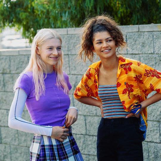 Euphoria character Jules Vaughn in long sleeves, with purple tee over and check skirt stands with another character, Rue Bennett, in orange and red Hawaiian shirt, striped tube top and black pants. 