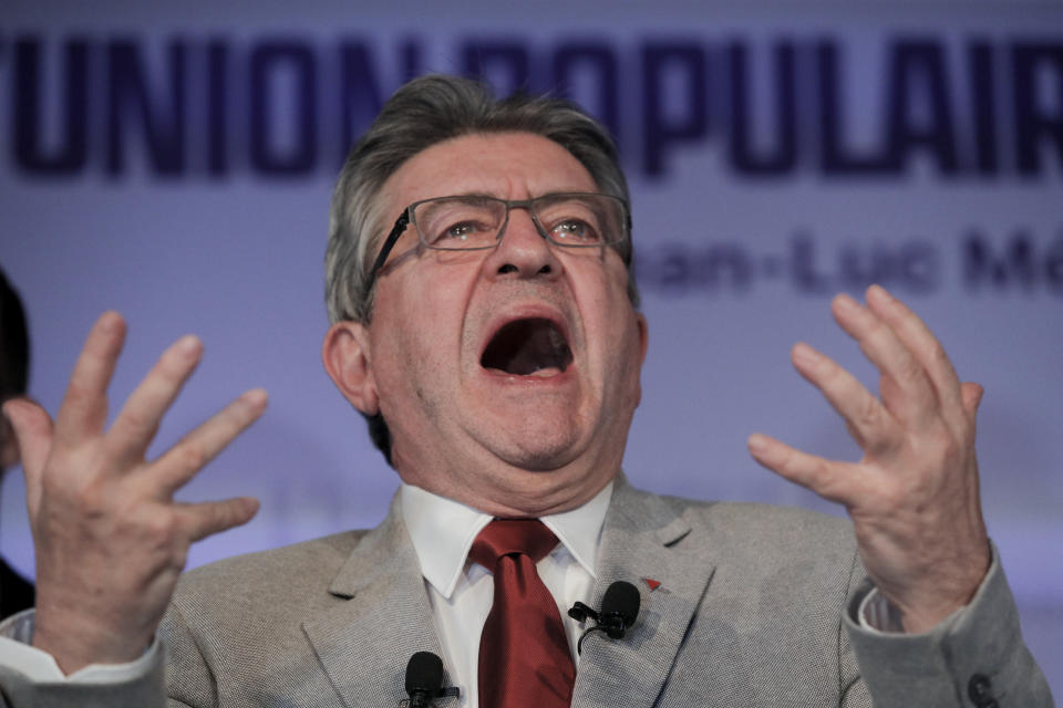 Jean-Luc Melenchon captured in full shout, with both hands gesturing boldly.