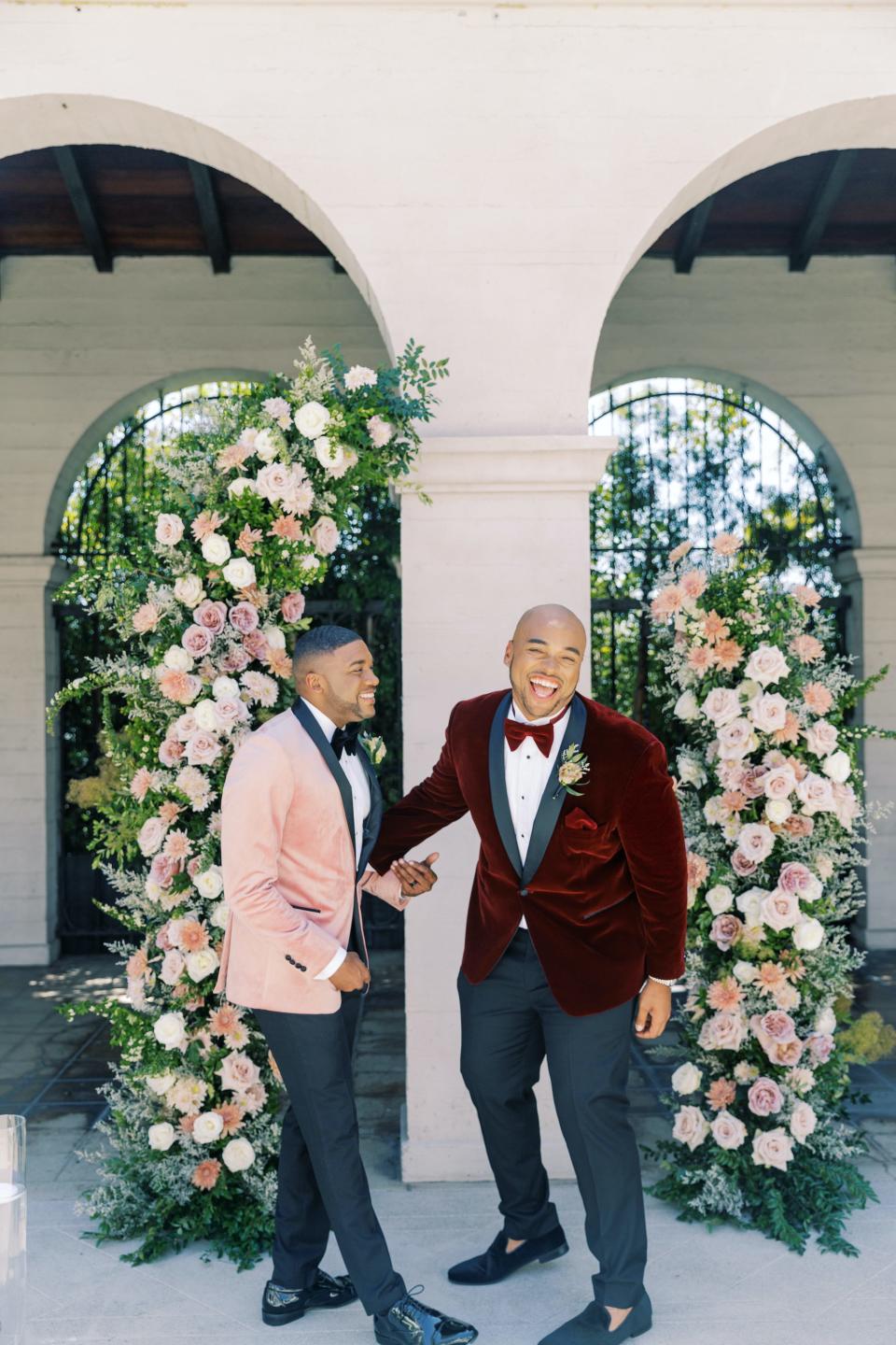 Two grooms wearing brightly colored suits laugh in front of flowers.