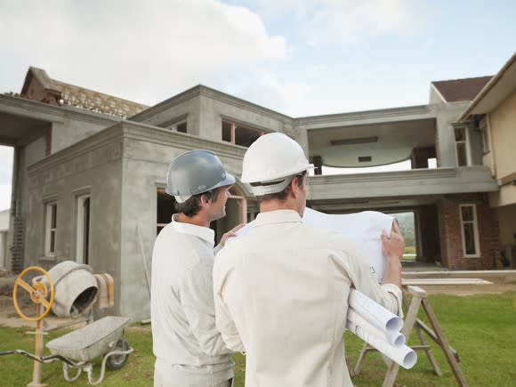 Two construction workers in hard hats facing a house while consulting house blueprints