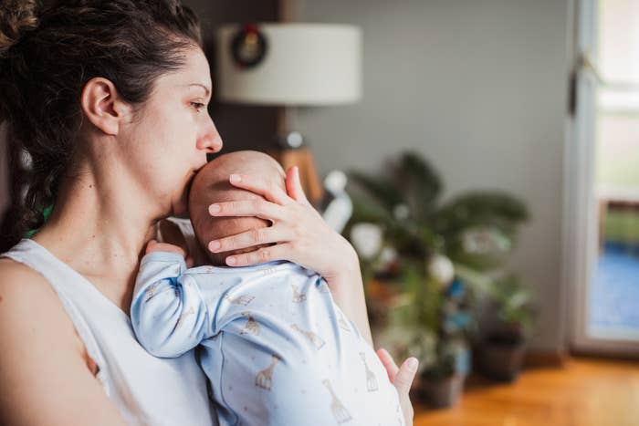 A woman holds a baby dressed in a patterned onesie closely to her chest, gently cradling the baby's head with her hand in a cozy indoor setting