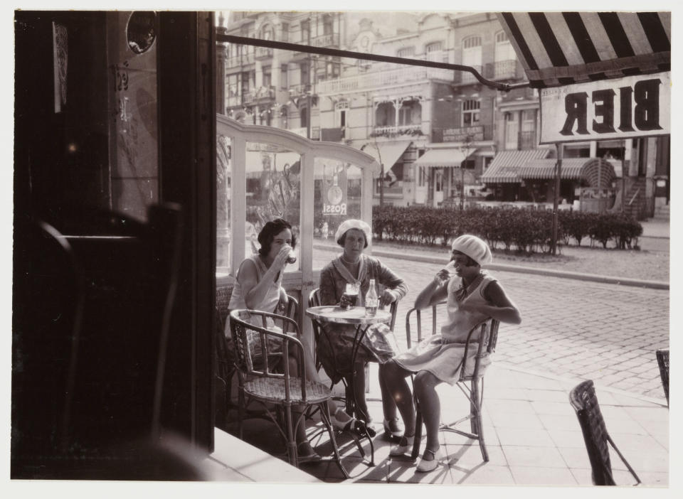 Three women in 1920s-style dresses and cloche hats sit at an outdoor café table, drinking and chatting