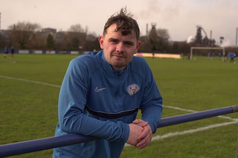 Man in blue top leans over barrier with sports field behind him