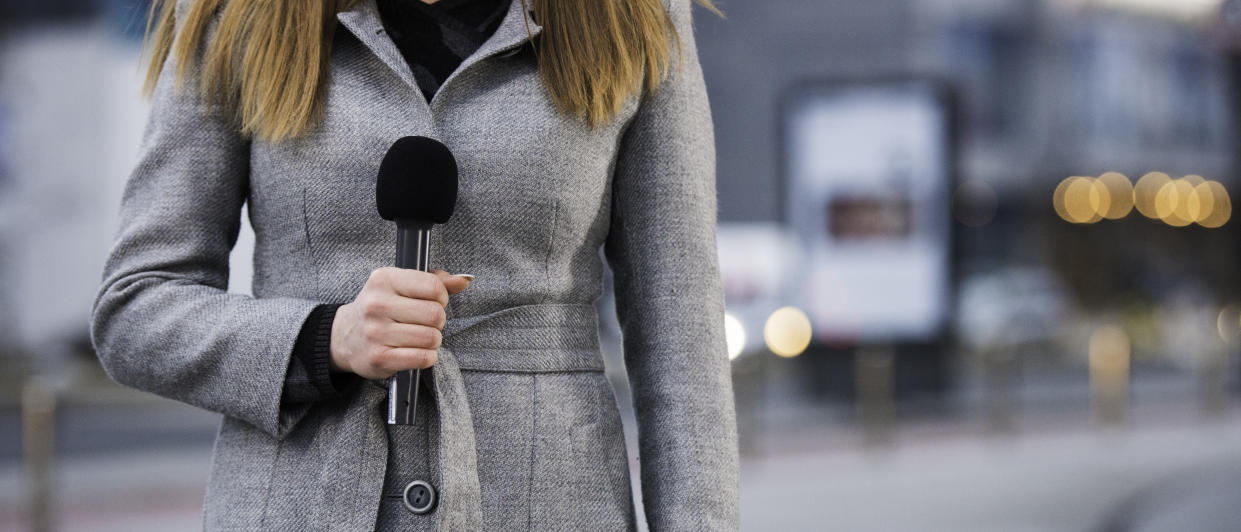 Front view of unrecognizable female  newscaster wearing coat and holding microphone, blurred city buildings in background, copy space.