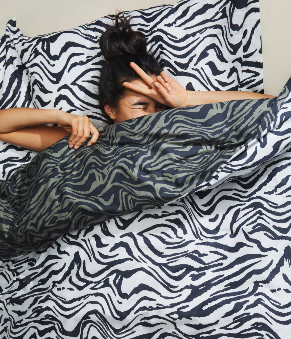 Woman snuggles in bed giving peace sign from zebra print reversible doona and pillow in white and navy and navy and green.