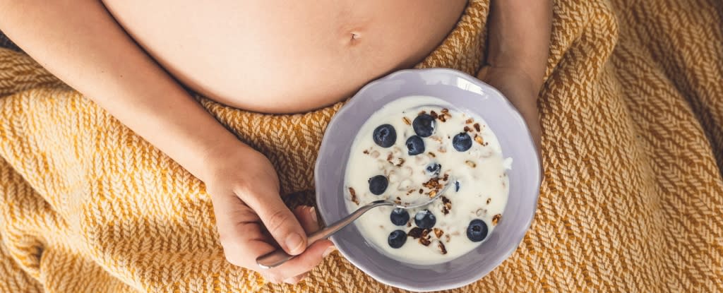 Hands holding a bowl of cereal and berries over a pregnant stomach partially covered by a yellow blanket