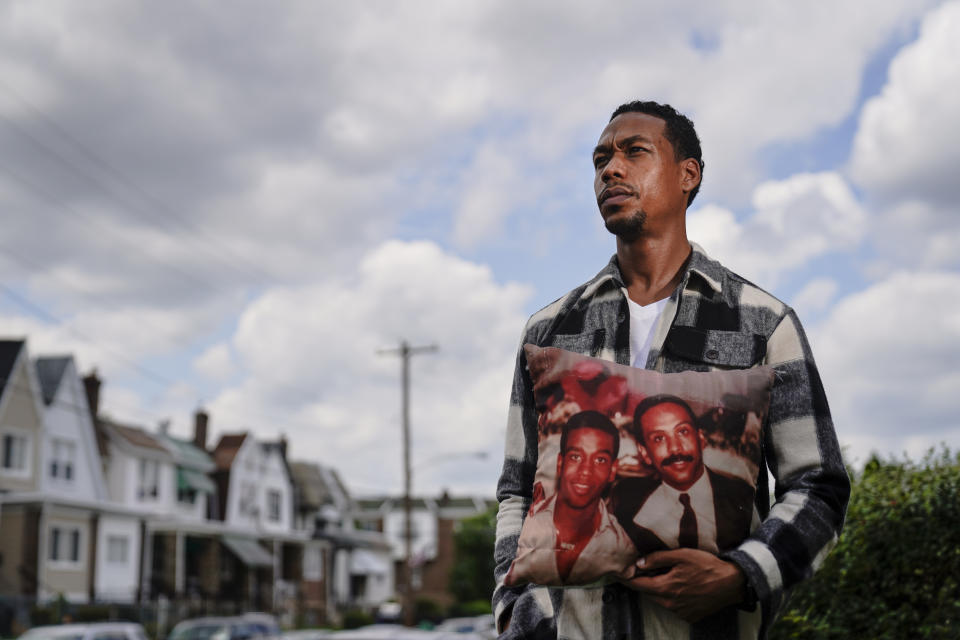 In this July 9, 2021, photo Brett Roman Williams poses for a photograph while holding a pillow with a photo of his father, Donald Williams, lower right, and brother Derrick Williams who both were killed by gunfire 20 years apart, in Philadelphia. (AP Photo/Matt Rourke)