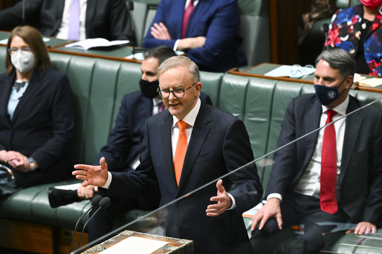 Australian Prime Minister Anthony Albanese stands at a podium in front of half a dozen others, who are seated on green-upholstered benches and wearing face masks.