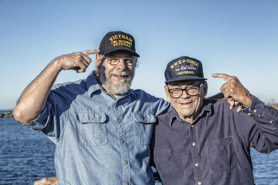 a vietnam and a korean war veteran pointing at their veteran hats