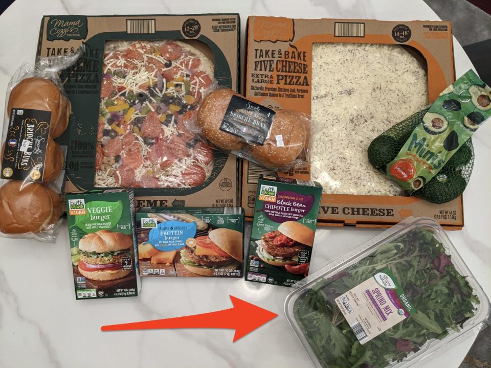 Pizza, broiche buns, avocafos, and veggies burgers all in an original packaging on a white table top as well as a red arrow pointing toward a plastic package of Aldi salad greens