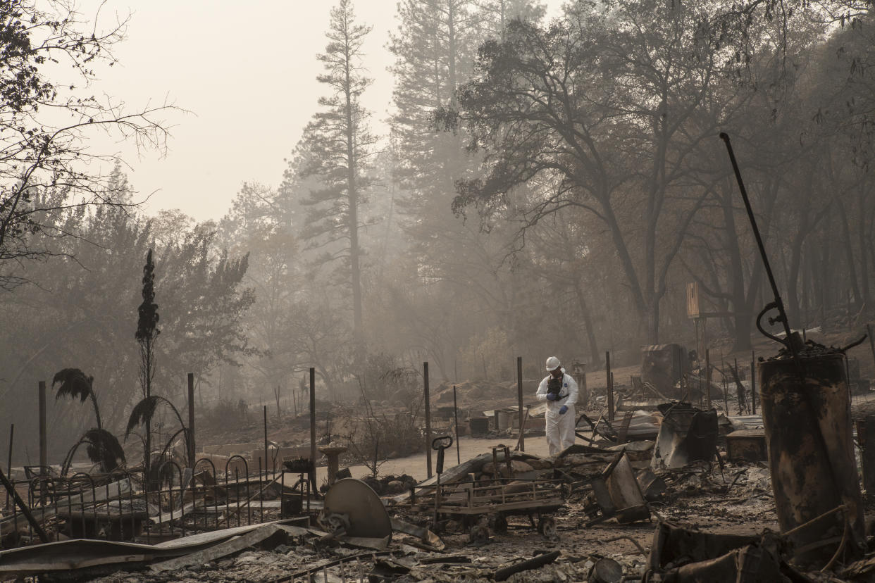 Person in white hazmat-type coveralls and white helmet stands looking down near the burned wreckage of where a home apparently once stood.