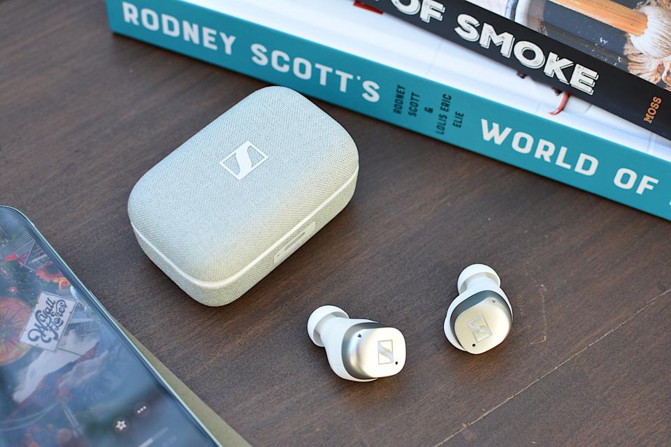 <p>White/silver Sennheiser Momentum True Wireless 4 earbuds laying on a wooden table with the case beside them.</p>
