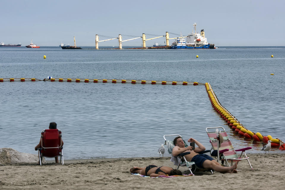 People relax on a beach in front of the stricken Tuvalu-registered OS 35 cargo ship that collided with a liquid natural gas carrier in the bay of Gibraltar last Monday in Gibraltar, Thursday, Sept. 1, 2022. Gibraltar authorities say that a small amount of heavy fuel oil has leaked from a bulk carrier ship stranded since colliding Monday with another ship near the Bay of Gibraltar. A government spokesman said that the situation was under control and the cargo ship was not in danger. He said there has been no environmental impact so far. (AP Photo/Marcos Moreno)