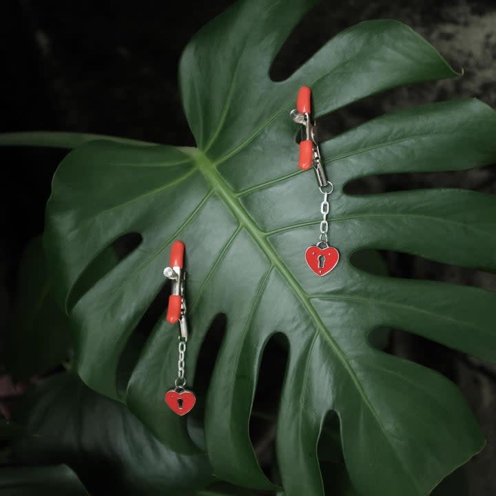Red nipple clamps with heart-shaped accent attached to monstera leaf