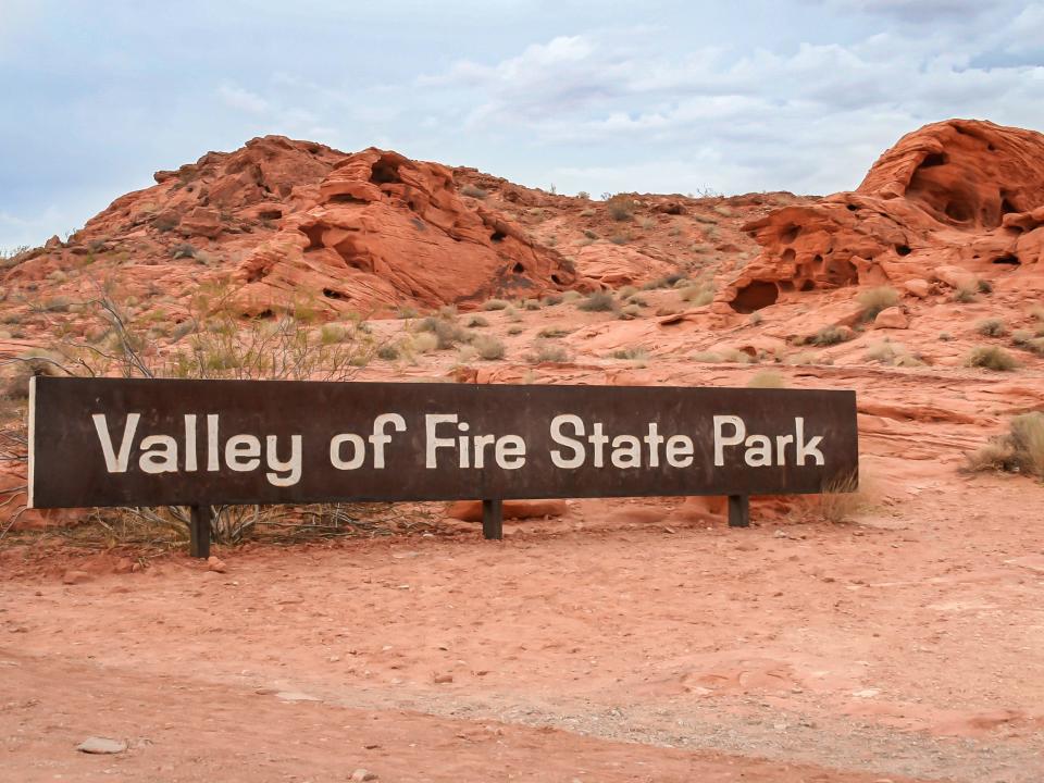 A long brown sign that reads "Valley of Fire State Park," with red-rock formations behind it.