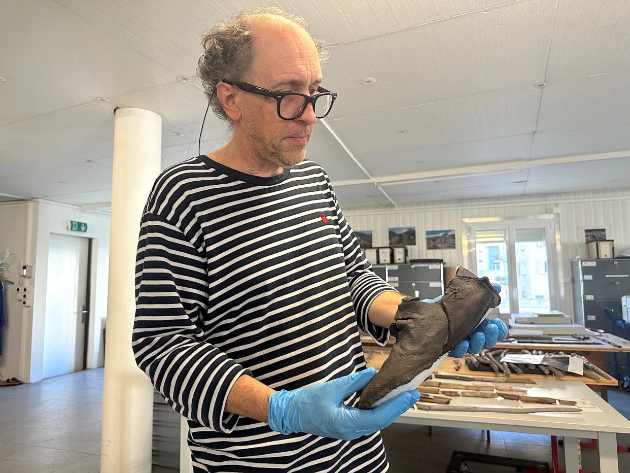 archaeologist with grey curly hair wearing glasses black and white striped t shirt and blue medical gloves holding a tattered ancient leather shoe