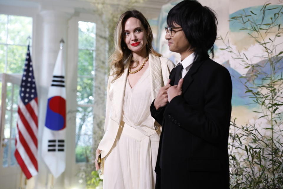 Two individuals standing next to flags, one in a white dress and the other in a suit