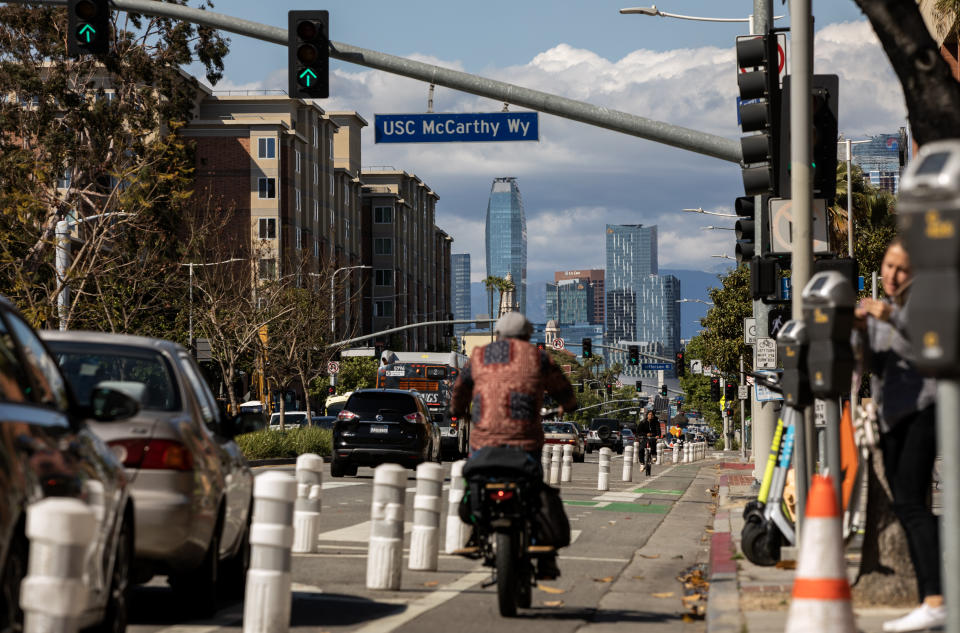 A motorcyclist, seen from behind, travels down a bike lane beneath a sign marked USC McCarthy Wy, as pedestrians wait at an intersection. Highrises can be seen in the distance.