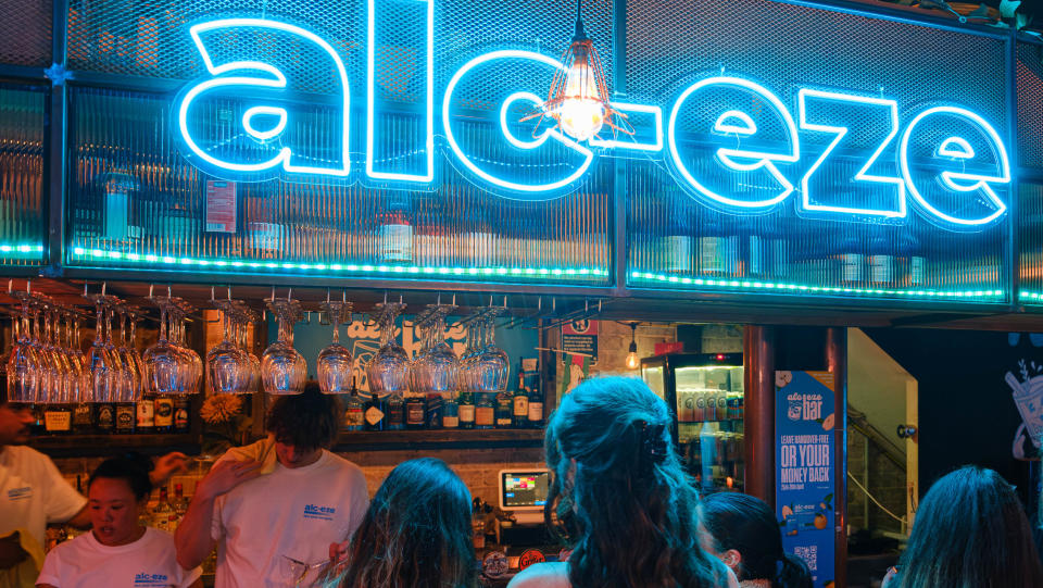 A blue neon sign over a bar with people standing at it. 